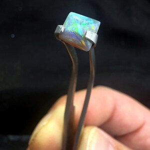 May include: A square-cut opal gemstone featuring a play of color with flashes of green, blue, and orange. The gemstone is held by a pair of silver-toned metal tweezers, showcasing its translucent quality and polished surface against a dark background.