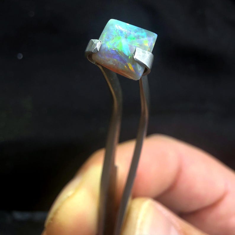 May include: A square-cut opal gemstone featuring a play of color with flashes of green, blue, and orange. The gemstone is held by a pair of silver-toned metal tweezers, showcasing its translucent quality and polished surface against a dark background.