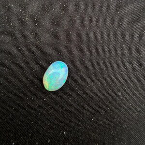 May include: An oval-shaped cabochon gemstone with a smooth, polished surface. The stone displays a play-of-color effect, featuring iridescent flashes of bright blue, green, and yellow against a translucent, milky white base color.