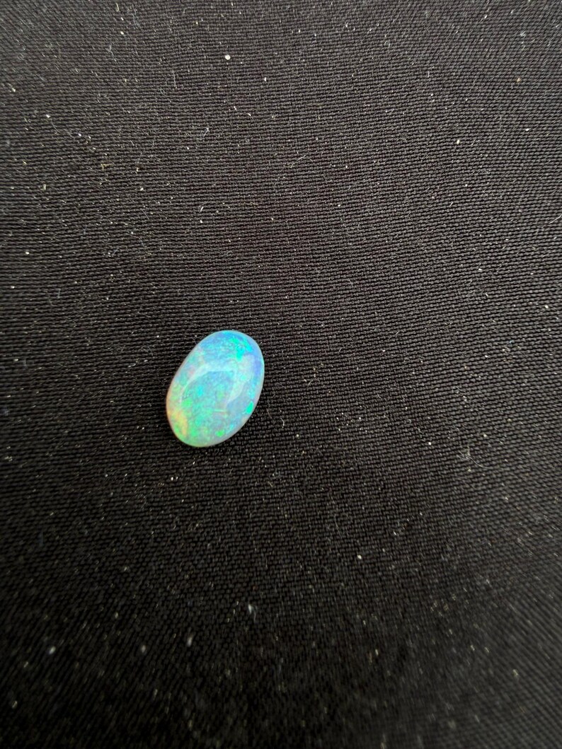 May include: An oval-shaped cabochon gemstone with a smooth, polished surface. The stone displays a play-of-color effect, featuring iridescent flashes of bright blue, green, and yellow against a translucent, milky white base color.