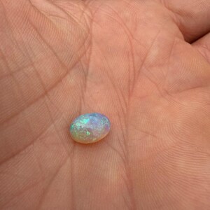 May include: An oval-shaped cabochon gemstone with a translucent, milky white base and vibrant flashes of iridescent green, blue, and pink light reflecting from within its polished, smooth, and rounded surface structure.