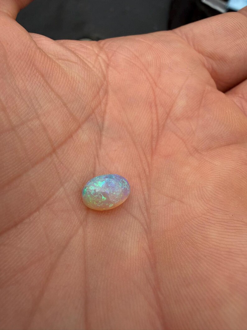 May include: An oval-shaped cabochon gemstone with a translucent, milky white base and vibrant flashes of iridescent green, blue, and pink light reflecting from within its polished, smooth, and rounded surface structure.