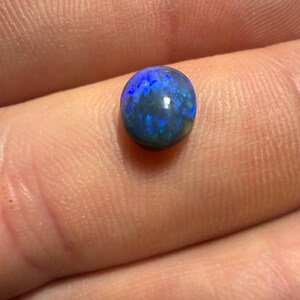 May include: A small, round, polished black opal gemstone featuring a dark base color with vibrant, iridescent flashes of electric blue and green light reflecting across its smooth, domed surface, showcasing a natural play of color.