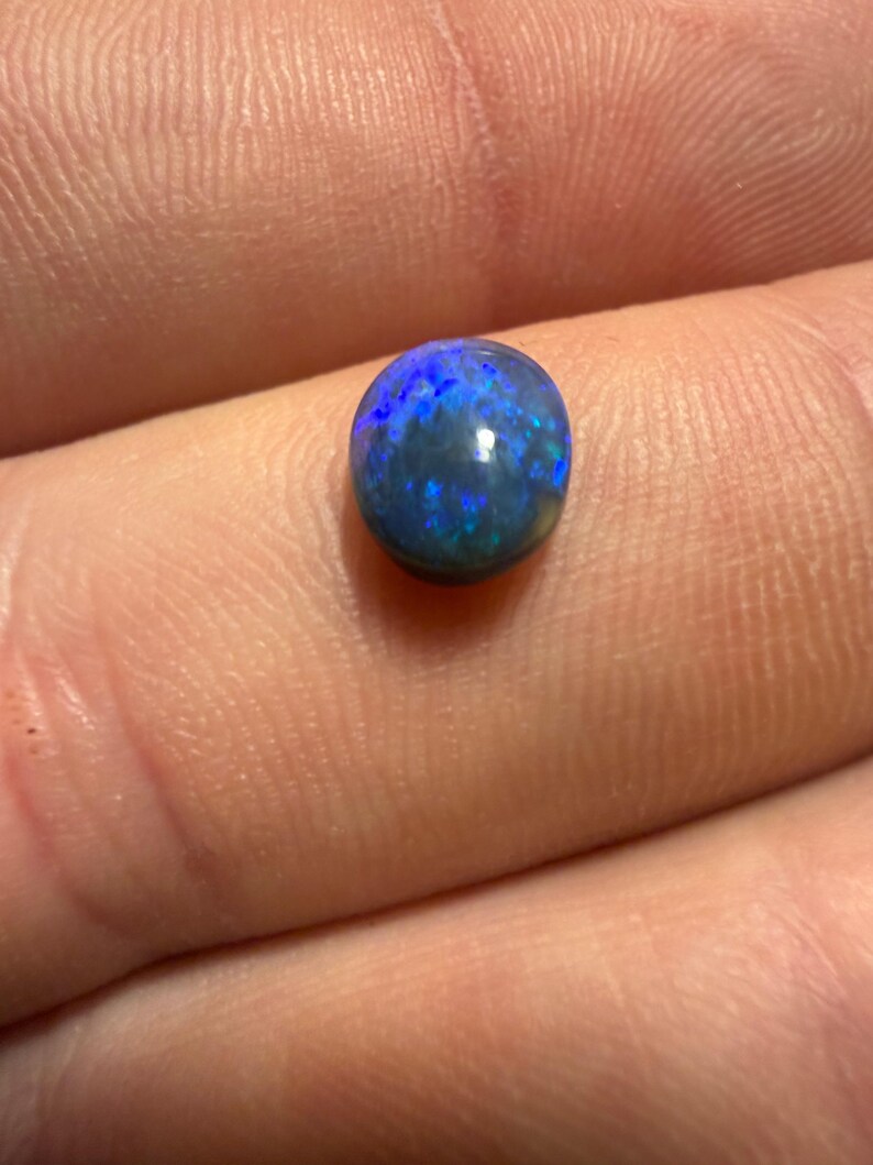May include: A small, round, polished black opal gemstone featuring a dark base color with vibrant, iridescent flashes of electric blue and green light reflecting across its smooth, domed surface, showcasing a natural play of color.