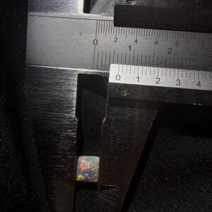 May include: A square-cut opal gemstone with a translucent base and vibrant multicolored flashes of red, green, and blue. The stone is positioned between the jaws of a metal caliper, showing a width of approximately 0.35 inches on the imperial scale.