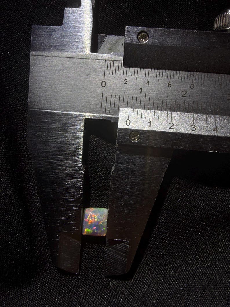 May include: A square-cut opal gemstone with a translucent base and vibrant multicolored flashes of red, green, and blue. The stone is positioned between the jaws of a metal caliper, showing a width of approximately 0.35 inches on the imperial scale.