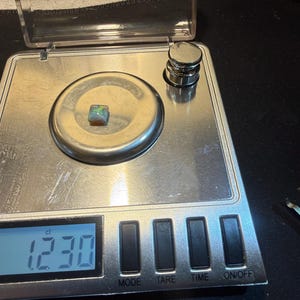 May include: A small, square-shaped opal gemstone with iridescent flashes of green, blue, and orange resting on a digital scale. The scale display shows a weight of 1.230 ct, and a small metal calibration weight is positioned in the upper right corner.