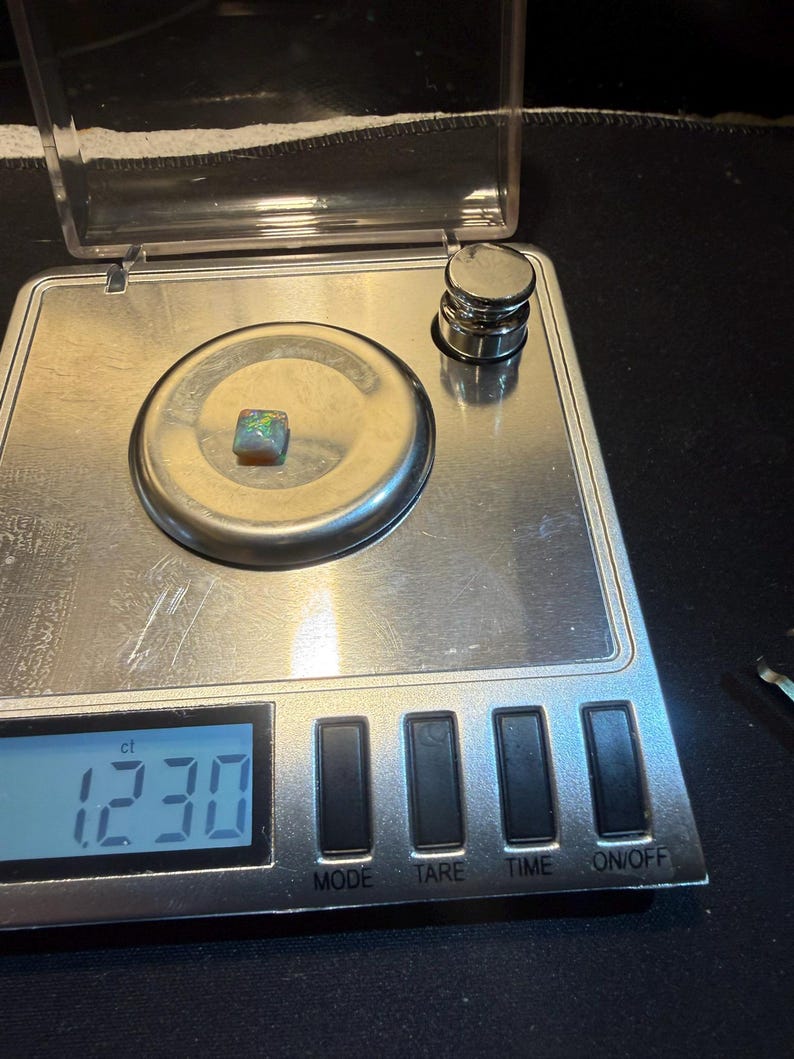 May include: A small, square-shaped opal gemstone with iridescent flashes of green, blue, and orange resting on a digital scale. The scale display shows a weight of 1.230 ct, and a small metal calibration weight is positioned in the upper right corner.