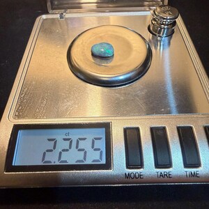 May include: A small, oval-shaped opal gemstone with iridescent green and blue flashes resting on a digital scale. The scale display clearly shows a weight of 2.255 ct, and a 20g calibration weight is visible in the corner of the metal platform.