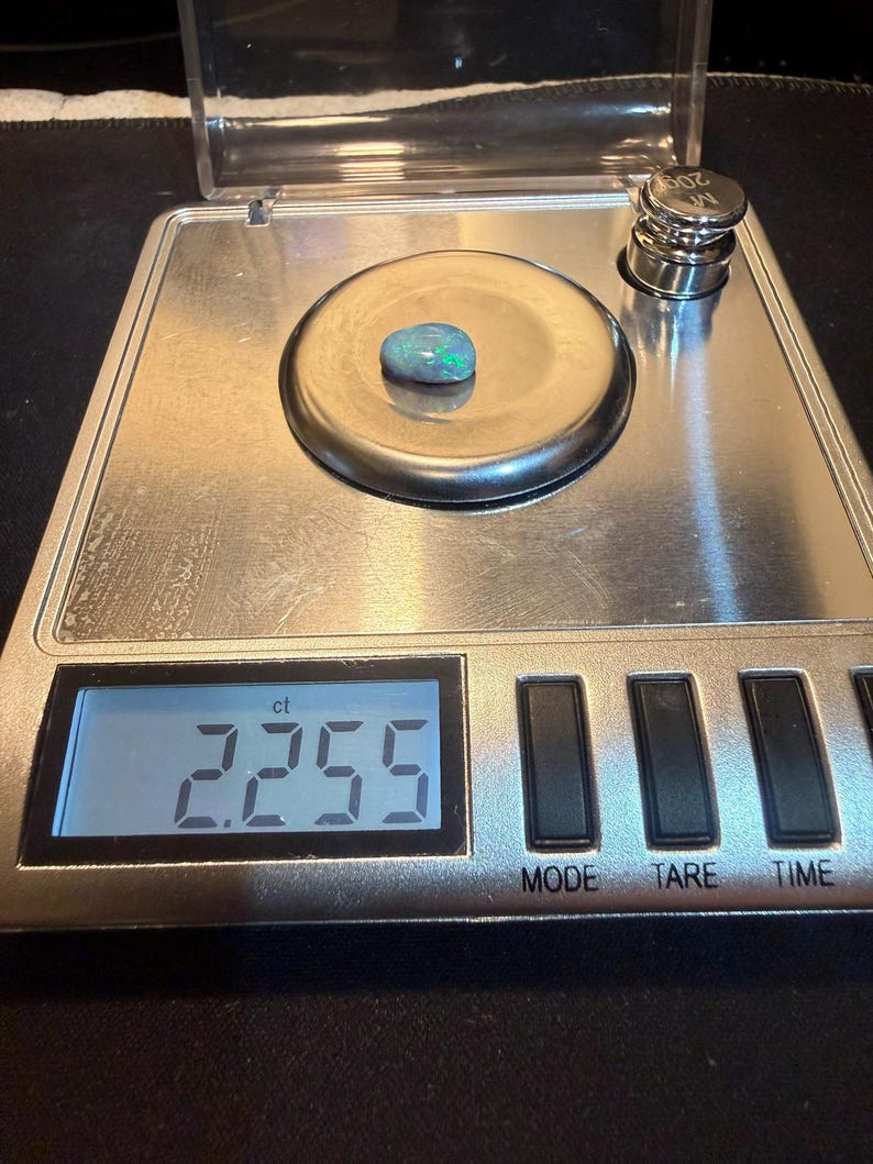 May include: A small, oval-shaped opal gemstone with iridescent green and blue flashes resting on a digital scale. The scale display clearly shows a weight of 2.255 ct, and a 20g calibration weight is visible in the corner of the metal platform.