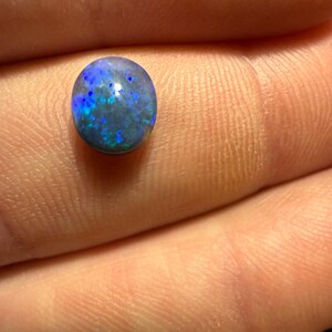 May include: A small, oval-shaped black opal gemstone featuring a dark base color with vibrant, iridescent flashes of blue and green light reflecting across its polished, smooth, and rounded surface.