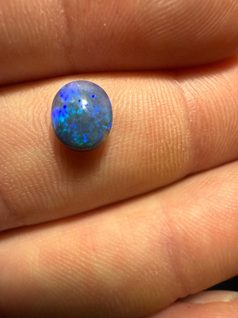 May include: A small, oval-shaped black opal gemstone featuring a dark base color with vibrant, iridescent flashes of blue and green light reflecting across its polished, smooth, and rounded surface.