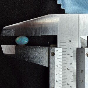 May include: An oval-shaped black opal gemstone with iridescent blue and green flashes of color. The gemstone is held within the jaws of a metal caliper, which indicates a width of approximately 0.35 inches on the imperial measurement scale.