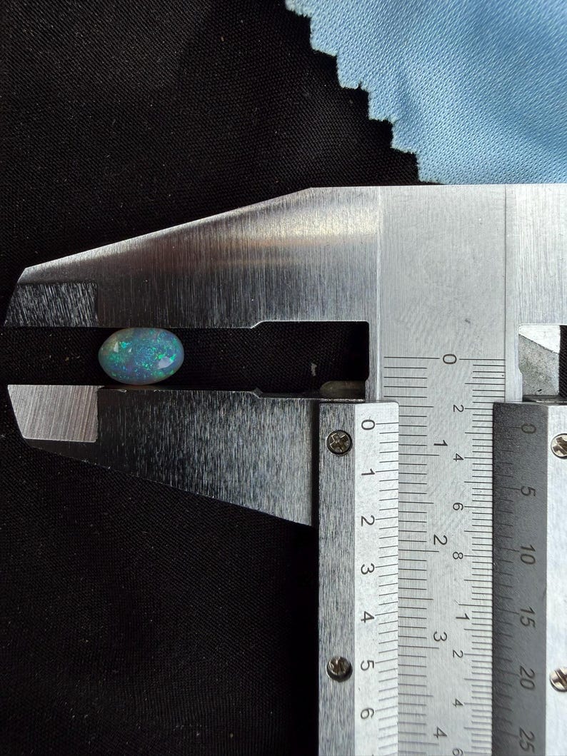 May include: An oval-shaped black opal gemstone with iridescent blue and green flashes of color. The gemstone is held within the jaws of a metal caliper, which indicates a width of approximately 0.35 inches on the imperial measurement scale.