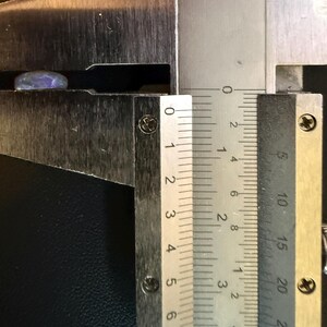 Może przedstawiać: Mały, opalizujący, owalny kamień szlachetny opal z niebieskimi i fioletowymi refleksami, umieszczony między szczękami metalowej suwmiarki. Narzędzie pomiarowe wskazuje, że kamień ma szerokość około 9 milimetr&oacute;w.