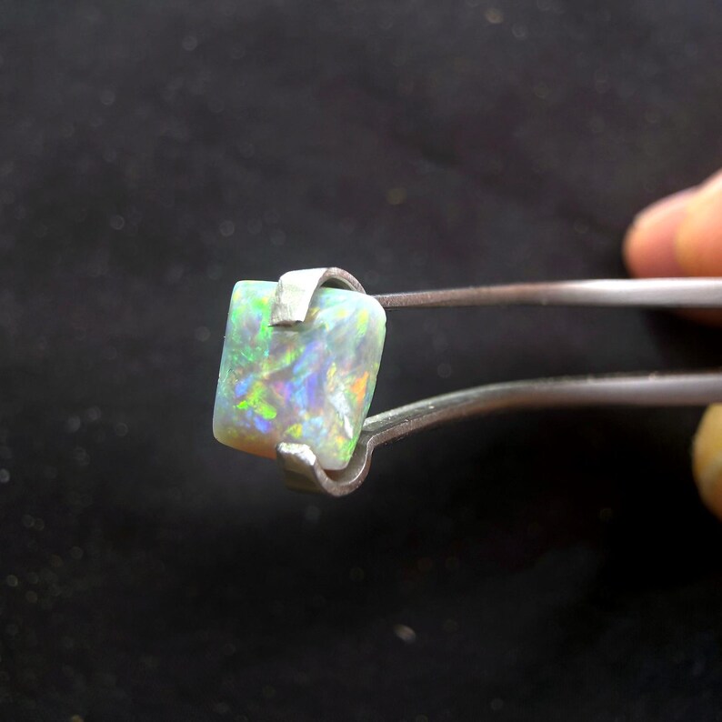 May include: A square-cut opal gemstone displaying a play of color with flashes of green, blue, and orange. The stone is held by a pair of metal tweezers, showcasing its translucent white base and vibrant internal light reflections.