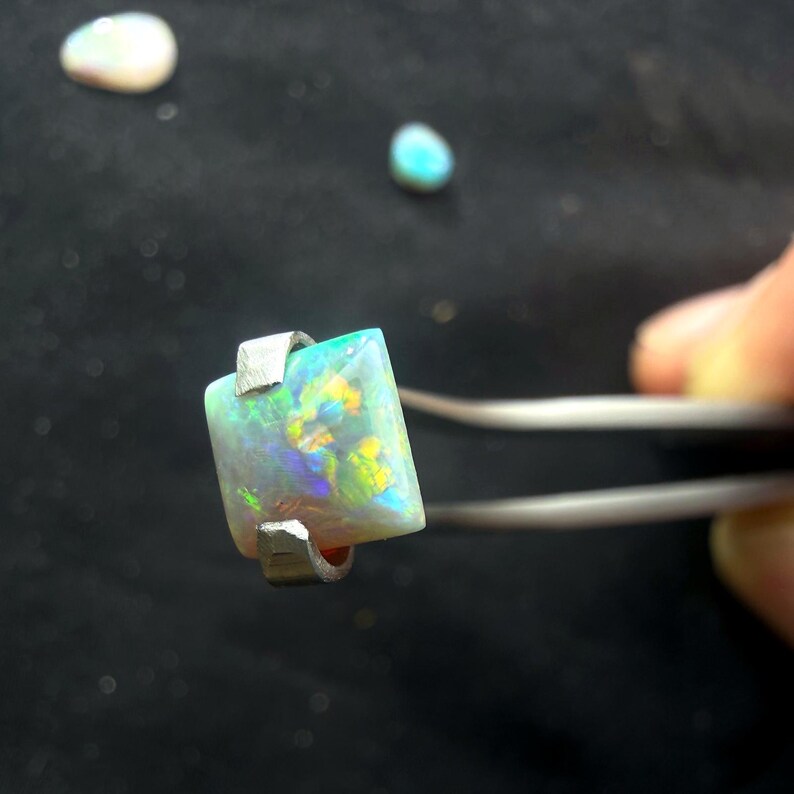 May include: A square-cut opal gemstone with a play-of-color featuring flashes of green, blue, and yellow, held by silver-toned metal tweezers. Two additional smaller, rounded, iridescent opal stones are visible in the background.