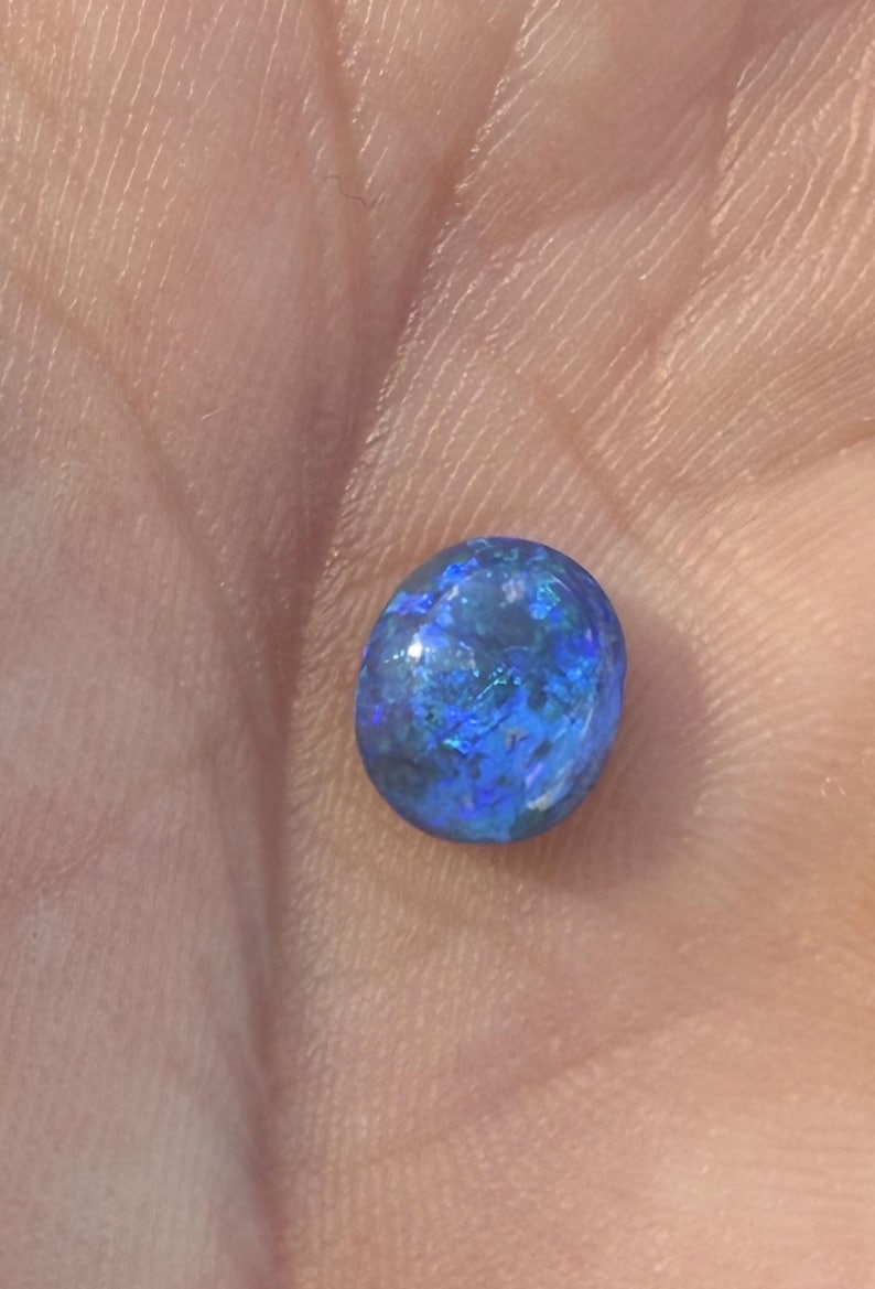 May include: A small, oval-shaped gemstone with a smooth, polished surface. The stone displays a deep, vibrant blue base color with iridescent flashes of lighter blue and hints of green throughout its translucent, crystalline structure.