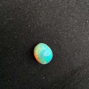 Peut inclure: Une pierre pr&eacute;cieuse opale de forme ovale lisse pr&eacute;sentant un jeu de couleurs vibrant avec des &eacute;clats iridescents de turquoise vif, de vert et de teintes orange chaud sur sa surface polie et arrondie, refl&eacute;tant la lumi&egrave;re avec &eacute;clat.