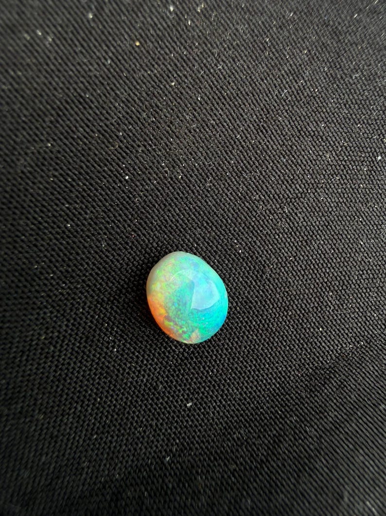 Peut inclure: Une pierre pr&eacute;cieuse opale de forme ovale lisse pr&eacute;sentant un jeu de couleurs vibrant avec des &eacute;clats iridescents de turquoise vif, de vert et de teintes orange chaud sur sa surface polie et arrondie, refl&eacute;tant la lumi&egrave;re avec &eacute;clat.
