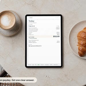 May include: A digital tablet displaying a financial planning application with a screen titled Today, showing a list of expenses, income, and a safe to spend balance of $312.00, alongside a cup of coffee and a croissant on a round table.