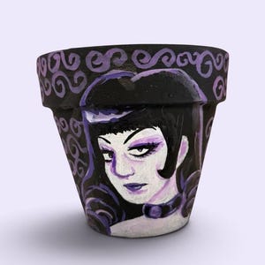 May include: A tapered terracotta plant pot painted in black and shades of purple. The exterior features a hand-painted portrait of a person with dark hair and purple makeup, surrounded by swirling decorative patterns on the rim and background.