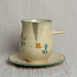 May include: A cream-colored ceramic creamer pitcher and matching saucer set. The pitcher features a small pouring spout, a side handle, and hand-painted floral motifs in orange and green. The saucer has a matching green rim and floral accents.