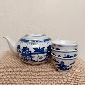 May include: A white ceramic teapot and three matching tea cups decorated with a blue willow pattern featuring pagodas, trees, and landscape elements. The teapot has a rounded body, a curved spout, and a lid, while the cups are stackable.