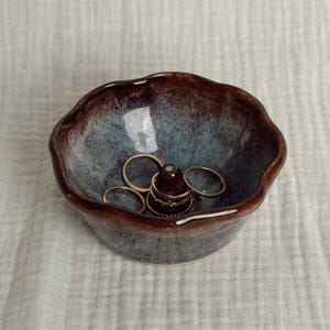 May include: A small, round, ceramic ring dish with a wavy, scalloped rim. The bowl features a reactive glaze with a blend of deep brown and mottled blue-grey tones. A central, conical post holds three thin, gold-colored metal rings.