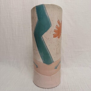 May include: A cylindrical ceramic vase with a speckled off-white upper section featuring a large teal zigzag stripe and an orange sun-like shape. The lower portion is a solid light pink with embossed geometric lines and small teal arrows.