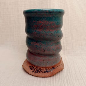 May include: A cylindrical ceramic tumbler with a wavy, ribbed exterior surface. The glaze features a mottled blend of teal and reddish-brown tones. The base is made of unglazed, light brown clay and features a dark, handwritten signature in black ink.