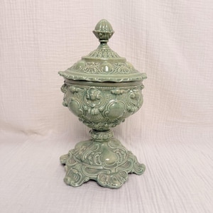 May include: A sage green ceramic lidded jar featuring an ornate, raised relief pattern across the body, lid, and base. The vessel has a pedestal foot, a rounded bowl, and a pointed finial on the lid, all finished in a glossy, monochromatic glaze.