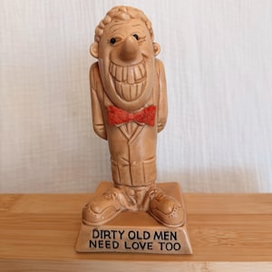 May include: A tan-colored figurine of a smiling character wearing a suit and a bright orange bow tie. The character stands on a rectangular base with the text "DIRTY OLD MEN NEED LOVE TOO" printed in black capital letters on the front face.