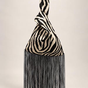 May include: A triangular-shaped handbag featuring a black and cream zebra stripe pattern. The bag has a knotted top handle and a long, straight fringe trim in solid black hanging from the bottom edge of the fabric body.