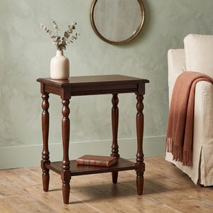 May include: A dark brown wooden side table with a rectangular top and a lower shelf. The table features four turned legs with decorative detailing. A light-colored ceramic vase with dried stems sits on the top, and a book rests on the lower shelf.