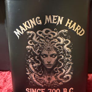 May include: A matte black metal flask featuring a graphic of Medusa with snakes for hair. The text "MAKING MEN HARD" is printed in an arched font above the image, and "SINCE 700 B.C." is printed in a straight font below the graphic.
