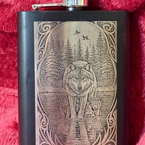 Könnte beinhalten: Ein schwarzer Metallflachmann mit einer silberfarbenen Lasergravur eines Wolfes, der mit zwei Welpen im Wasser steht. Das Motiv zeigt einen Wald im Hintergrund, Vögel am Himmel und einen verzierten Rahmen. Der Flachmann hat einen silbernen Schraubverschluss.