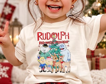 Puede incluir: Una camiseta de manga corta color crema con un estampado gráfico de los personajes de Rudolph el reno de la nariz roja, incluyendo un muñeco de nieve, un árbol de Navidad, un yeti, un elfo y renos, con el texto Rudolph the Red-Nosed Reindeer.