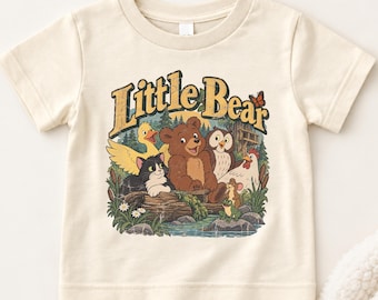 Puede incluir: Una camiseta de manga corta color crema con un estampado gráfico que incluye el texto Little Bear. La ilustración muestra un oso pardo, un gato blanco y negro, un pato amarillo, un búho, una gallina, un ratón y una rana en un bosque.