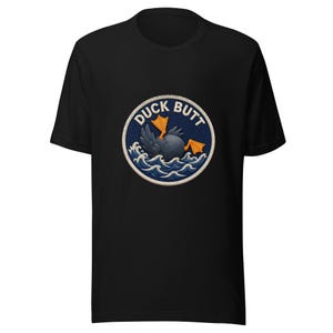 May include: A black short-sleeve t-shirt featuring a circular graphic on the chest. The design shows a dark gray duck diving into blue ocean waves with its orange webbed feet sticking out, surrounded by the text DUCK BUTT in white letters.