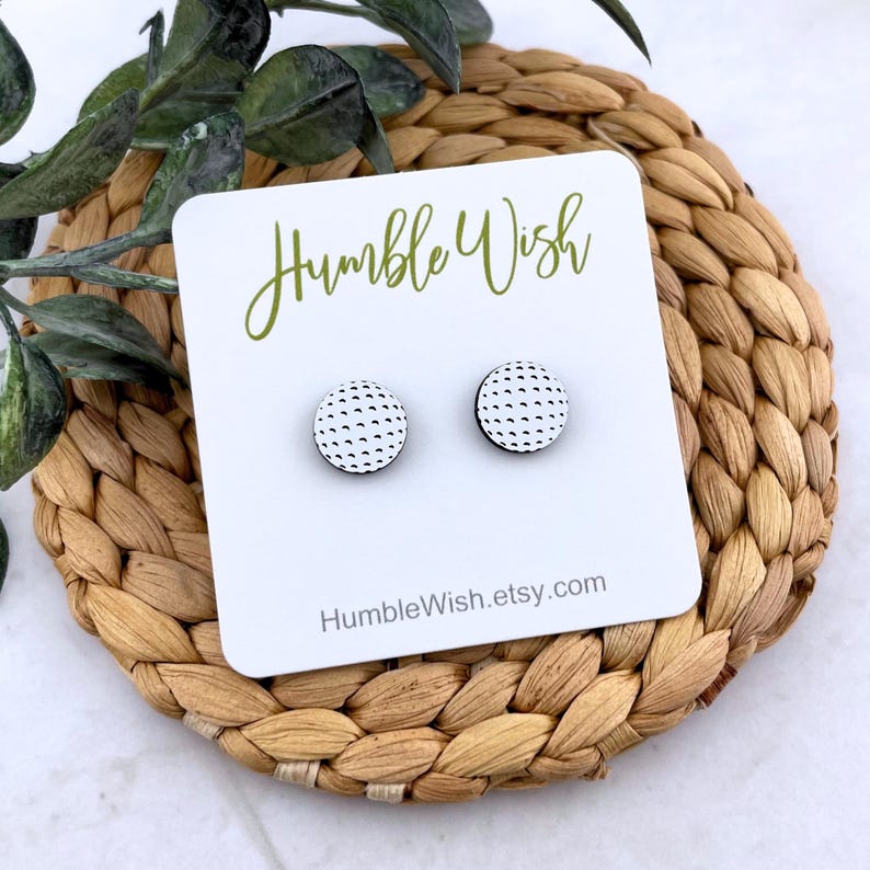 Peut inclure: Une paire de boucles d'oreilles rondes avec un motif de balle de golf. Les boucles d'oreilles sont blanches avec un motif &agrave; pois noirs. Elles sont pr&eacute;sent&eacute;es sur une carte blanche avec le texte "Humble Wish" et l'adresse du site web.