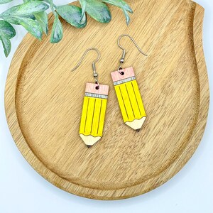 Peut inclure: Une paire de boucles d'oreilles en forme de crayon jaune avec des pointes d'effaceur roses et des crochets argentés.