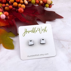 Peut inclure: Boucles d'oreilles fantômes blanches avec des yeux noirs et un contour noir, présentées sur une carte blanche. La carte porte l'inscription "Humble Wish" en vert et une adresse de site web. Décor automnal en arrière-plan.