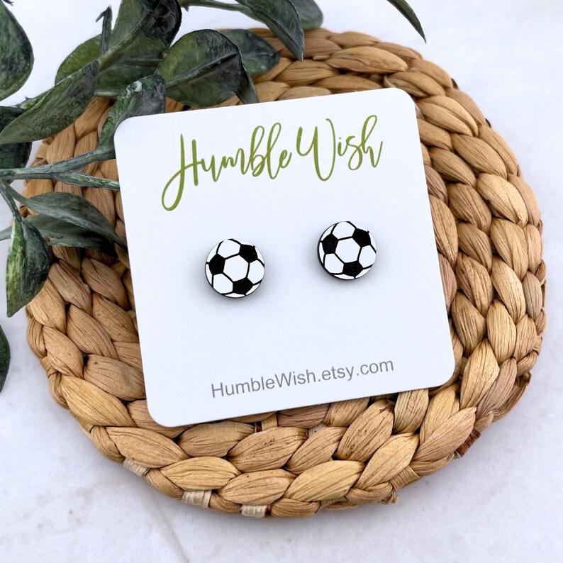 Peut inclure: Deux boucles d'oreilles &agrave; clous en forme de ballon de football noir et blanc sur une carte blanche avec le texte "Humble Wish" et l'adresse du site web. Les boucles d'oreilles sont pr&eacute;sent&eacute;es sur un dessous de plat circulaire tress&eacute;.