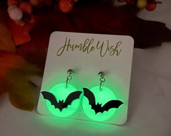 May include: A pair of glow-in-the-dark circular earrings featuring black bat silhouettes on a bright green background. The earrings are attached to a white display card with the text Humble Wish printed in a dark green script font.