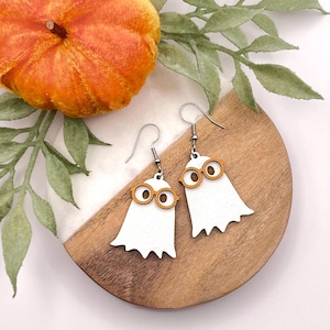 May include: A pair of white ghost earrings with orange glasses. The earrings are made of wood and have silver hooks.