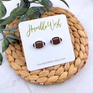 Peut inclure: Une paire de boucles d'oreilles &agrave; clous en forme de ballon de football. Les boucles d'oreilles sont marron avec des d&eacute;tails de couture blancs. Elles sont pr&eacute;sent&eacute;es sur une carte blanche avec le texte "Humble Wish" en &eacute;criture verte et l'adresse du site Web "HumbleWish.etsy.com".