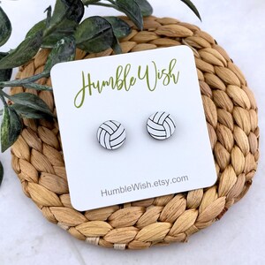 Peut inclure: Deux boucles d'oreilles clous en forme de ballon de volley-ball sur une carte blanche avec le texte "Humble Wish" en vert. Les boucles d'oreilles sont blanches avec des lignes noires. La carte repose sur un dessous de plat tress&eacute;.