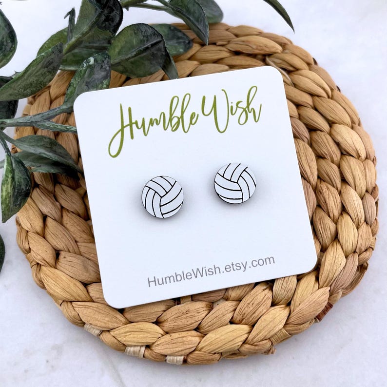 Peut inclure: Deux boucles d'oreilles clous en forme de ballon de volley-ball sur une carte blanche avec le texte "Humble Wish" en vert. Les boucles d'oreilles sont blanches avec des lignes noires. La carte repose sur un dessous de plat tress&eacute;.