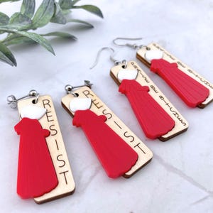 May include: A pair of wooden earrings with a red handmaid's tale inspired design. The earrings feature a red silhouette of a woman in a red robe with a white bonnet. The earrings are engraved with the word "RESIST".
